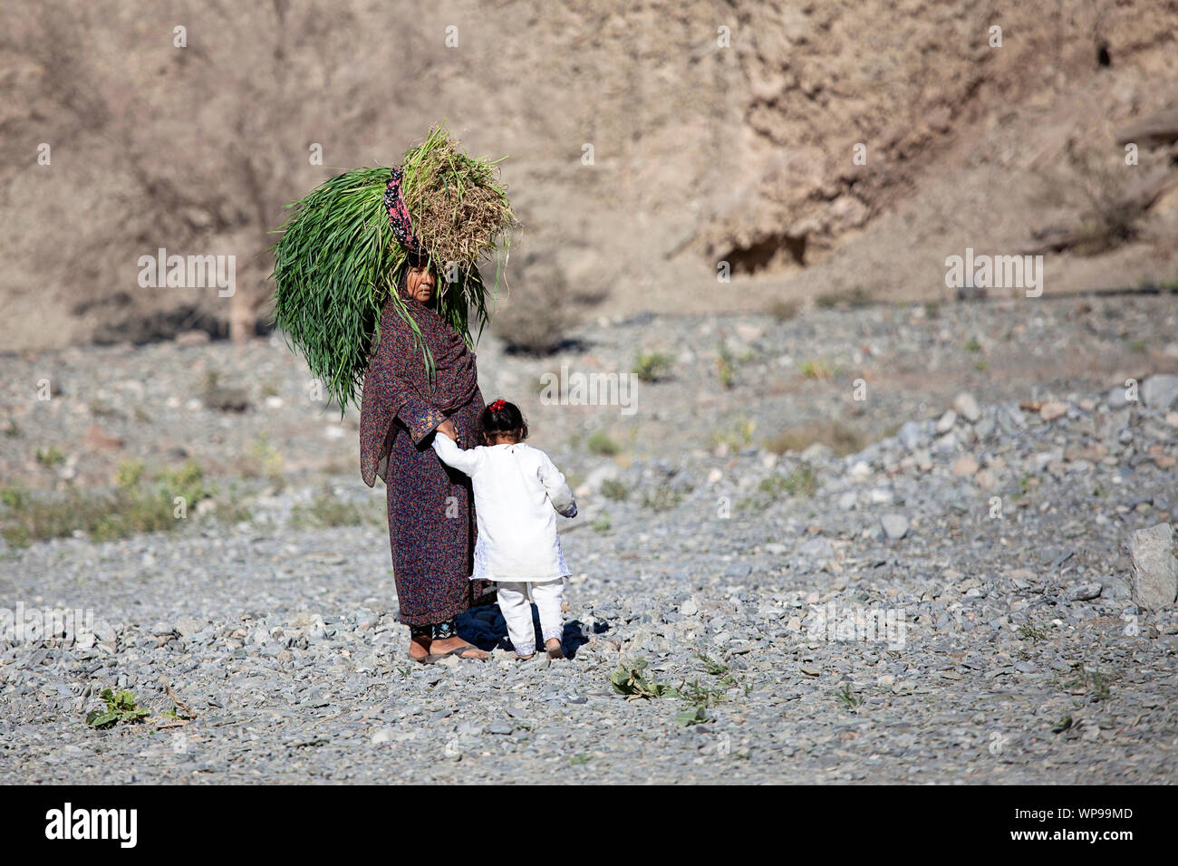 Frau trägt an Kopf große Scheiben von Heu und halten ein Kind in der ariden Landschaft im Oman Stockfoto