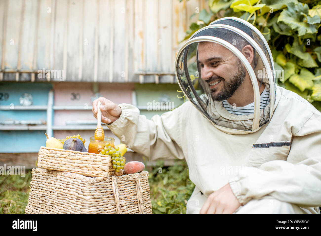 Porträt einer fröhlichen Imker in Schützende Uniform mit Honig und süßen Früchten auf die Imkerei mit Bienenstöcken auf dem Hintergrund Stockfoto