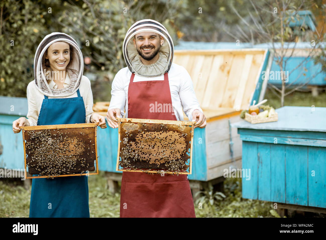 Porträt einer männlichen und weiblichen Imker in schützende Hüte und Schürzen holding Waben auf die Imkerei mit hölzernen Bienenstöcke auf dem Hintergrund Stockfoto