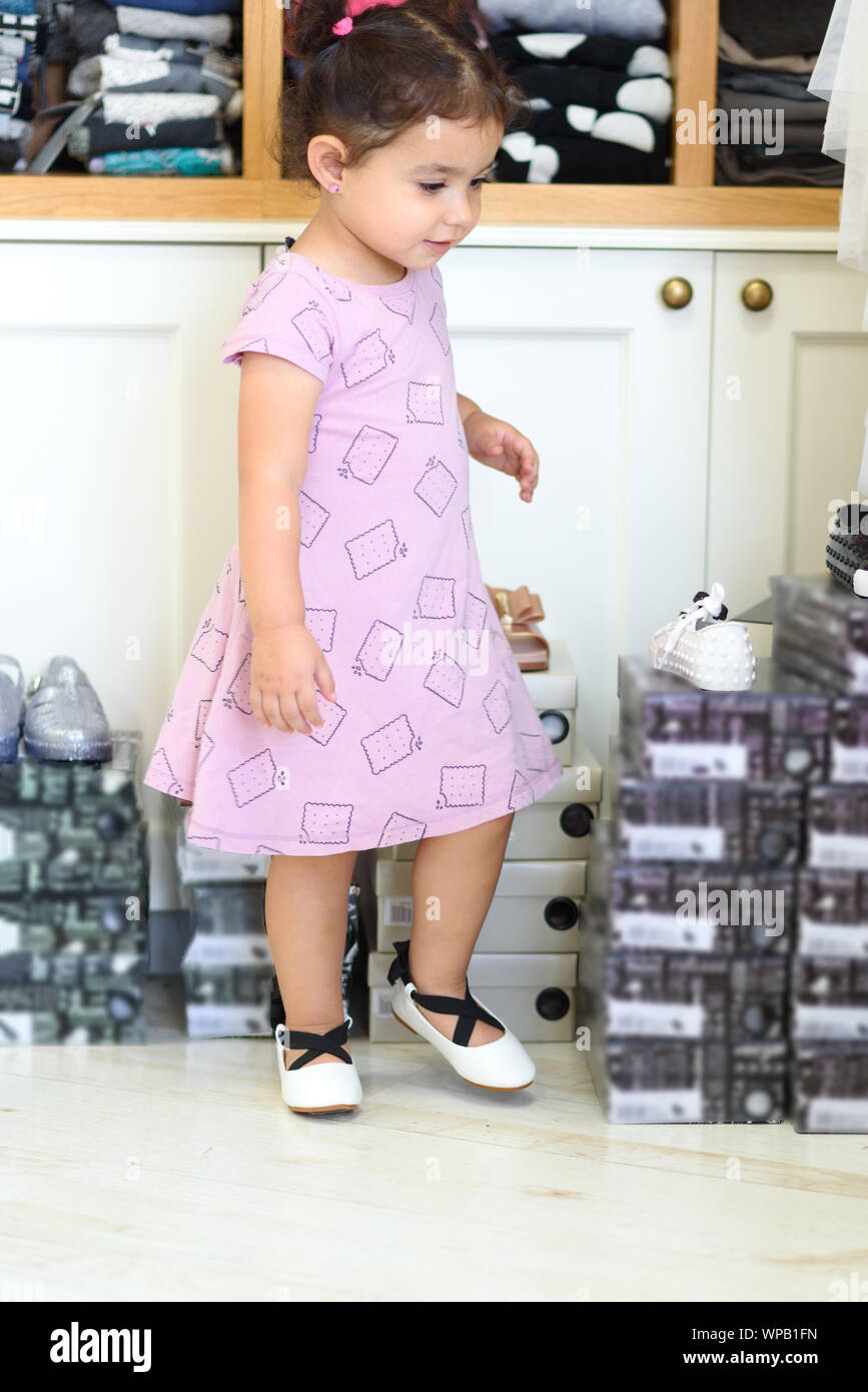 Süße kleine Mädchen in einem Schuh- und Kleidung Store mit neuen Kleid und  Wohnungen. Kleinkind Kind Mädchen spielen in einem Laden. Wenig  multirassischen Kind auswählen und versuchen, auf neue Schuhe und Kleidung