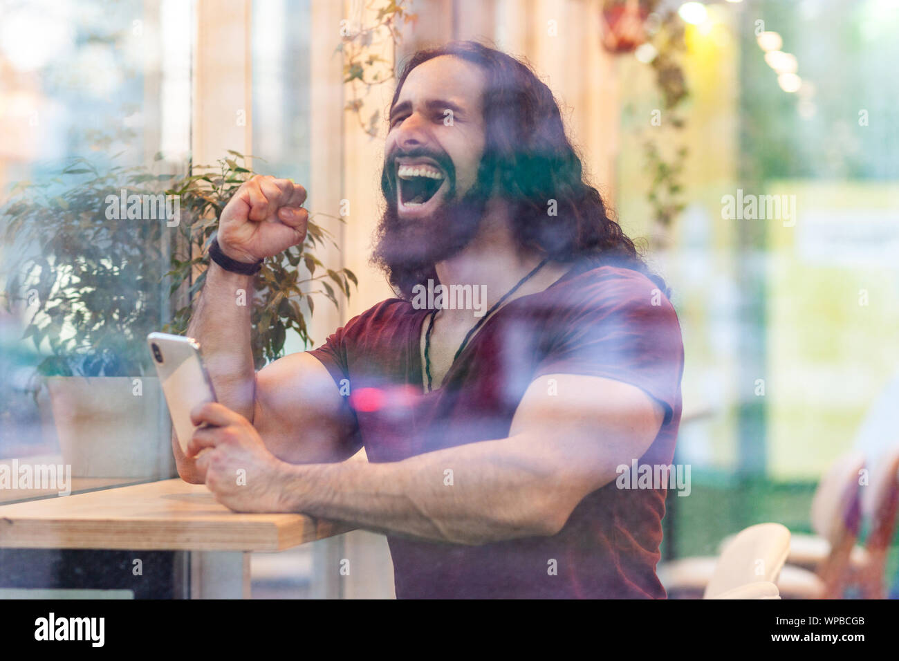 Seitenansicht Portrait durch das Fenster von zufriedenen hübscher junger Erwachsener sportliche Meister Mann mit lockigem Haar lang sitzen auf Cafe, Telefon und tr Stockfoto