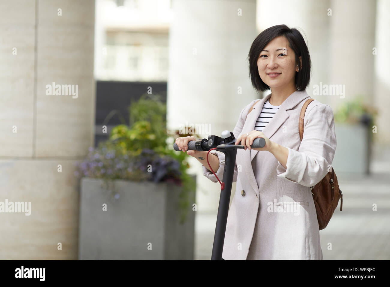Taille bis Portrait von lächelnden Asiatin, Elektroroller, Kamera, während in der Stadt Straße Posing, kopieren Raum Stockfoto