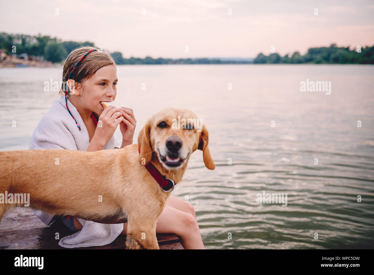 Mädchen sitzt auf einem Dock mit Ihrem Hund und essen Sandwich am Fluss Stockfoto