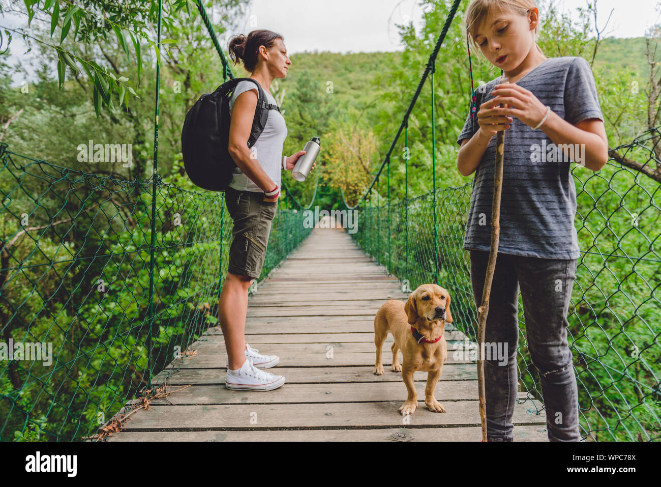 Mutter und Tochter stehen auf hölzerne Hängebrücke mit kleinen gelben Hund im Wald Stockfoto