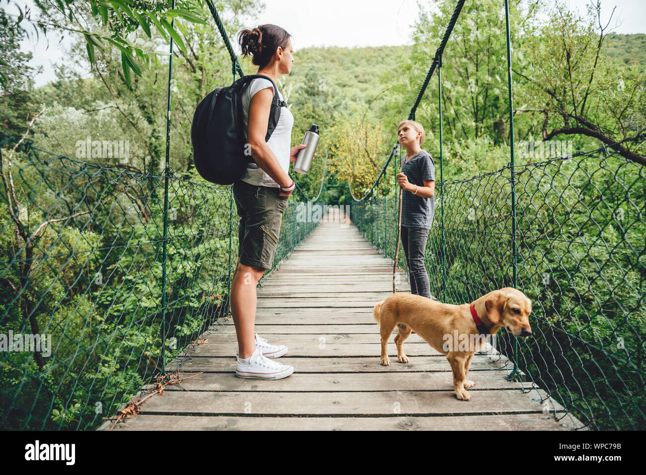 Mutter und Tochter stehen auf hölzerne Hängebrücke mit kleinen gelben Hund im Wald Stockfoto