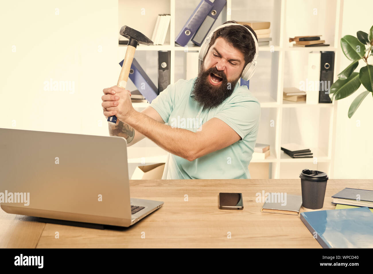 Veraltete Software. Computer lag. Gründe für Computer zurückbleiben. Wie fix langsam hinterher. Hasse office Routine. Man bärtige Kerl Kopfhörer Büro schwingen Hammer auf dem Computer. Langsame Internetverbindung. Stockfoto