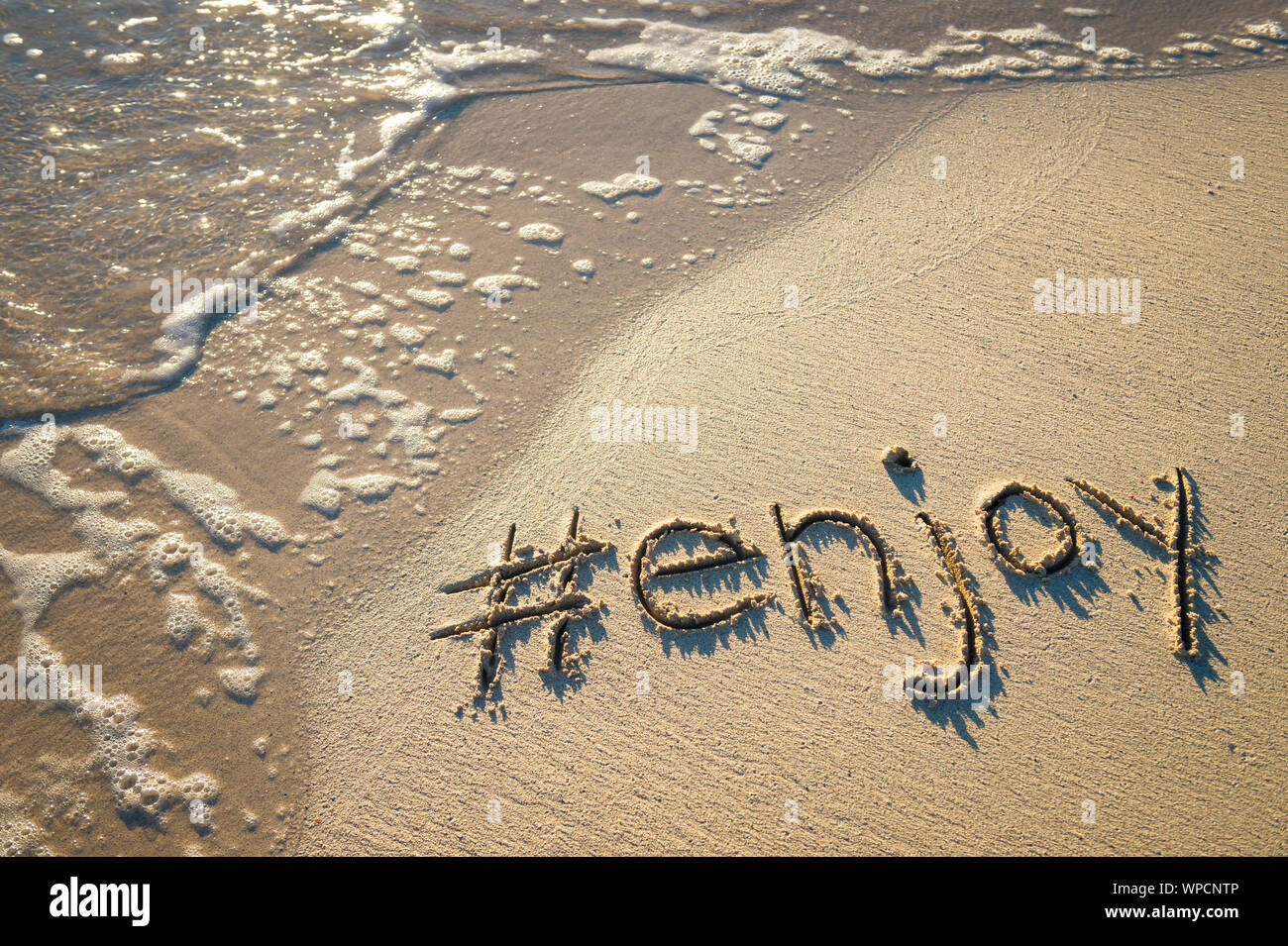 Modernes reisen Nachricht für den Strand 'genießen' mit einem Social Media - freundliche hashtag in glatten Sand mit ankommenden Welle geschrieben Stockfoto