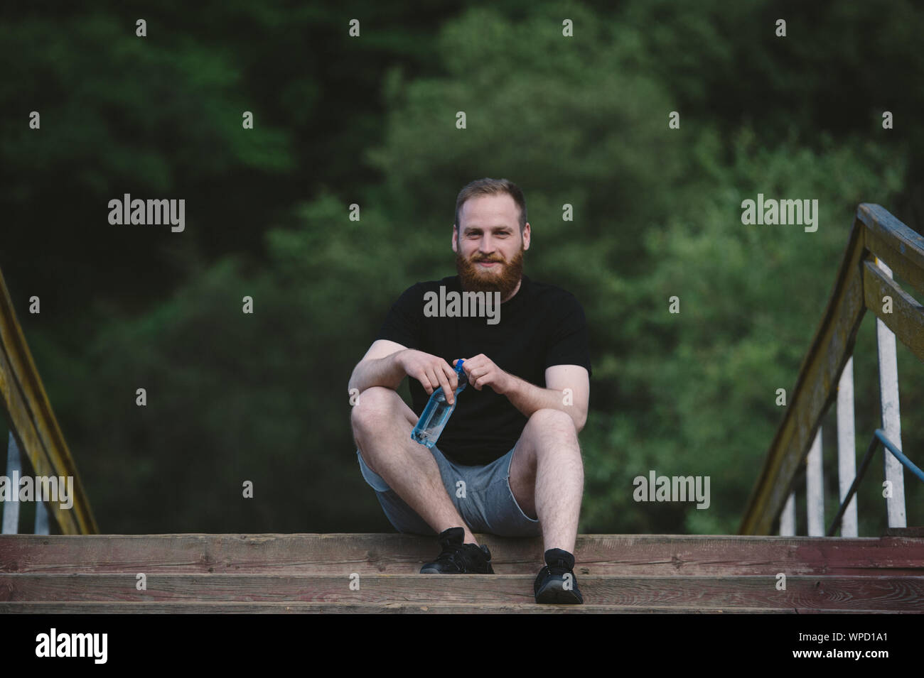 Gesunde junge bärtiger Mann im schwarzen T-Shirt und graue Shorts und Turnschuhen Sitzen im Freien auf Holzbrücke im Green Park während der Rest holding Flasche Stockfoto
