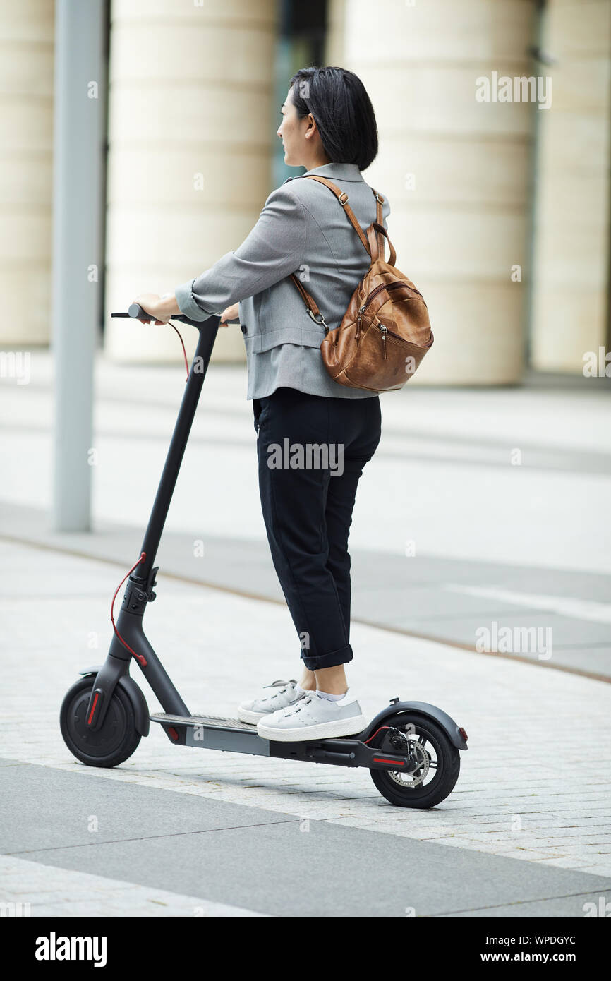 Volle Länge Seitenansicht des jungen asiatischen Frau reiten Elektroroller in Stadt Straße mit Architektur im Hintergrund, kopieren Raum Stockfoto