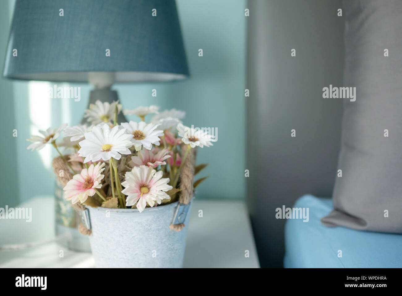 Nahaufnahme, Blumen und die Lampe auf dem Tisch mit Sunray im Schlafzimmer. Grau Kissen auf die blaue Bettdecke. Stockfoto