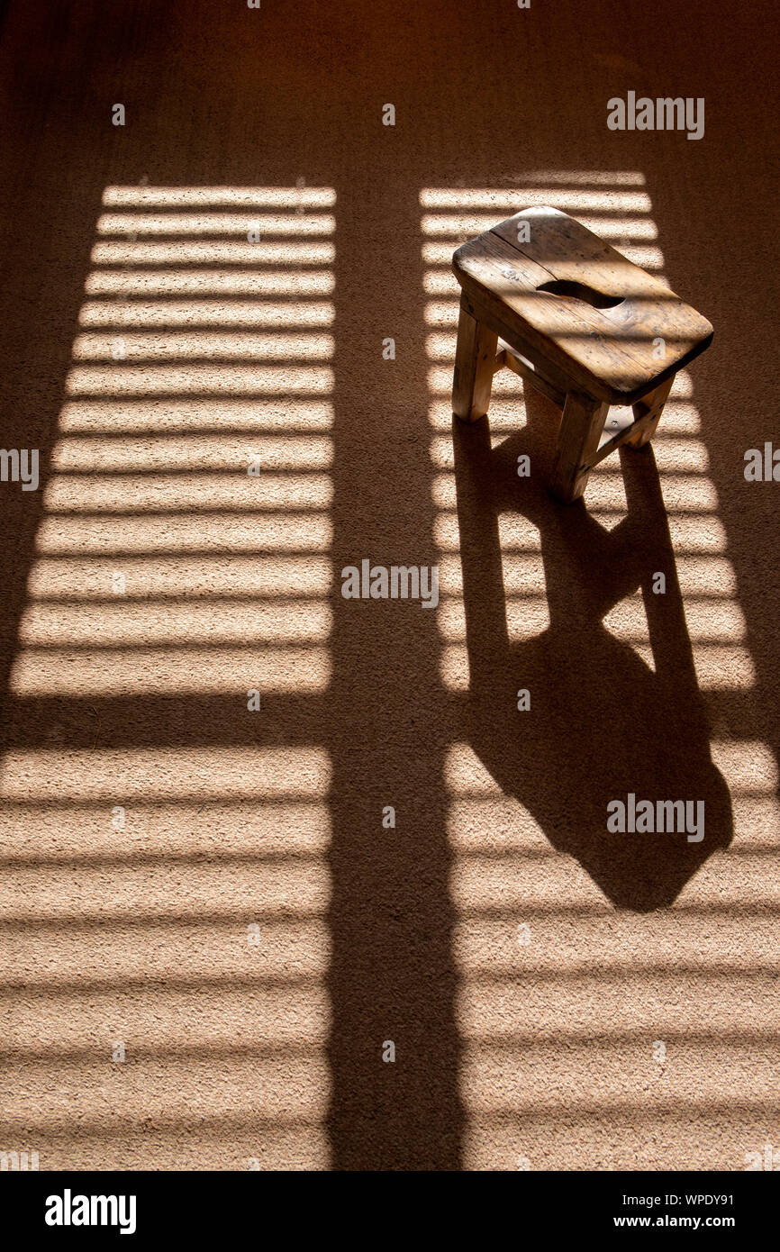 Sun peeping durch Fenster mit geschlossenen Jalousien und Melken Hocker mit Schatten spielen auf braunen Teppich Stockfoto