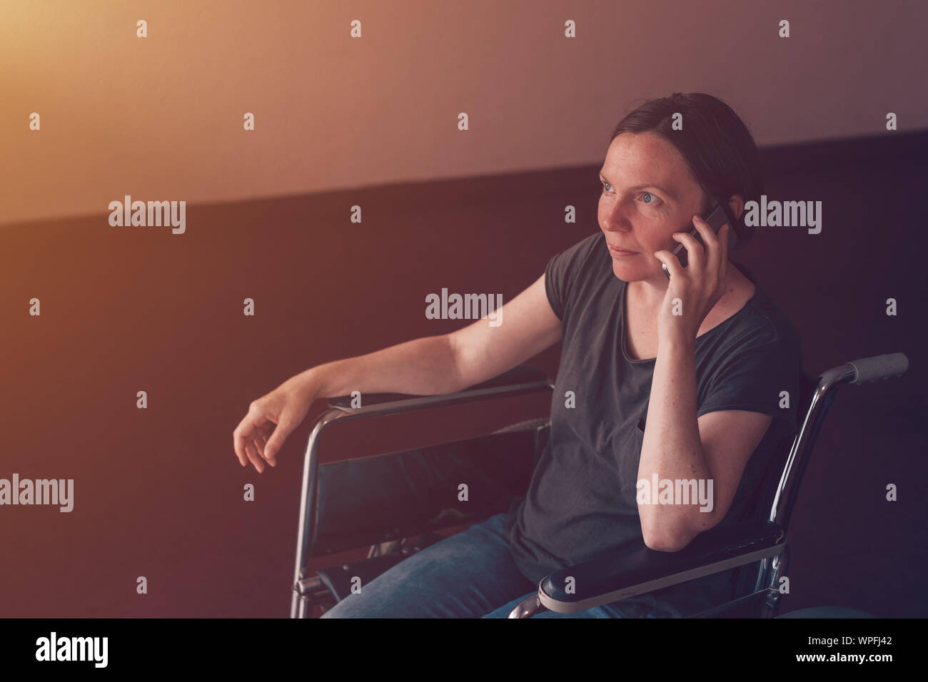 Hoffnungsvolle weibliche Person mit Behinderung im Rollstuhl Gespräch am Handy und schauen aus dem Fenster in Pflegeheim Stockfoto