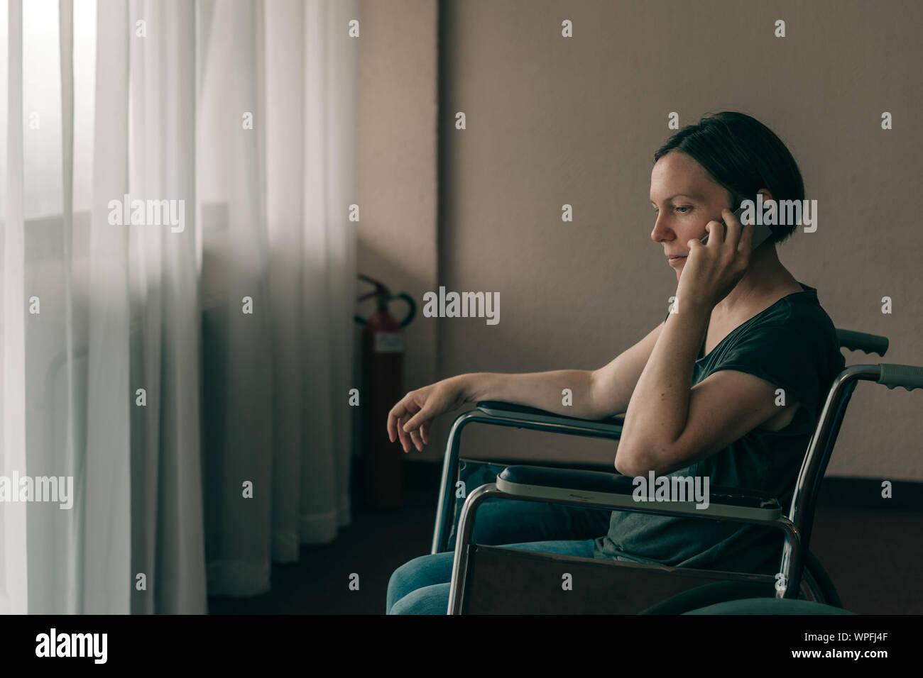 Hoffnungsvolle weibliche Person mit Behinderung im Rollstuhl Gespräch am Handy und schauen aus dem Fenster in Pflegeheim Stockfoto