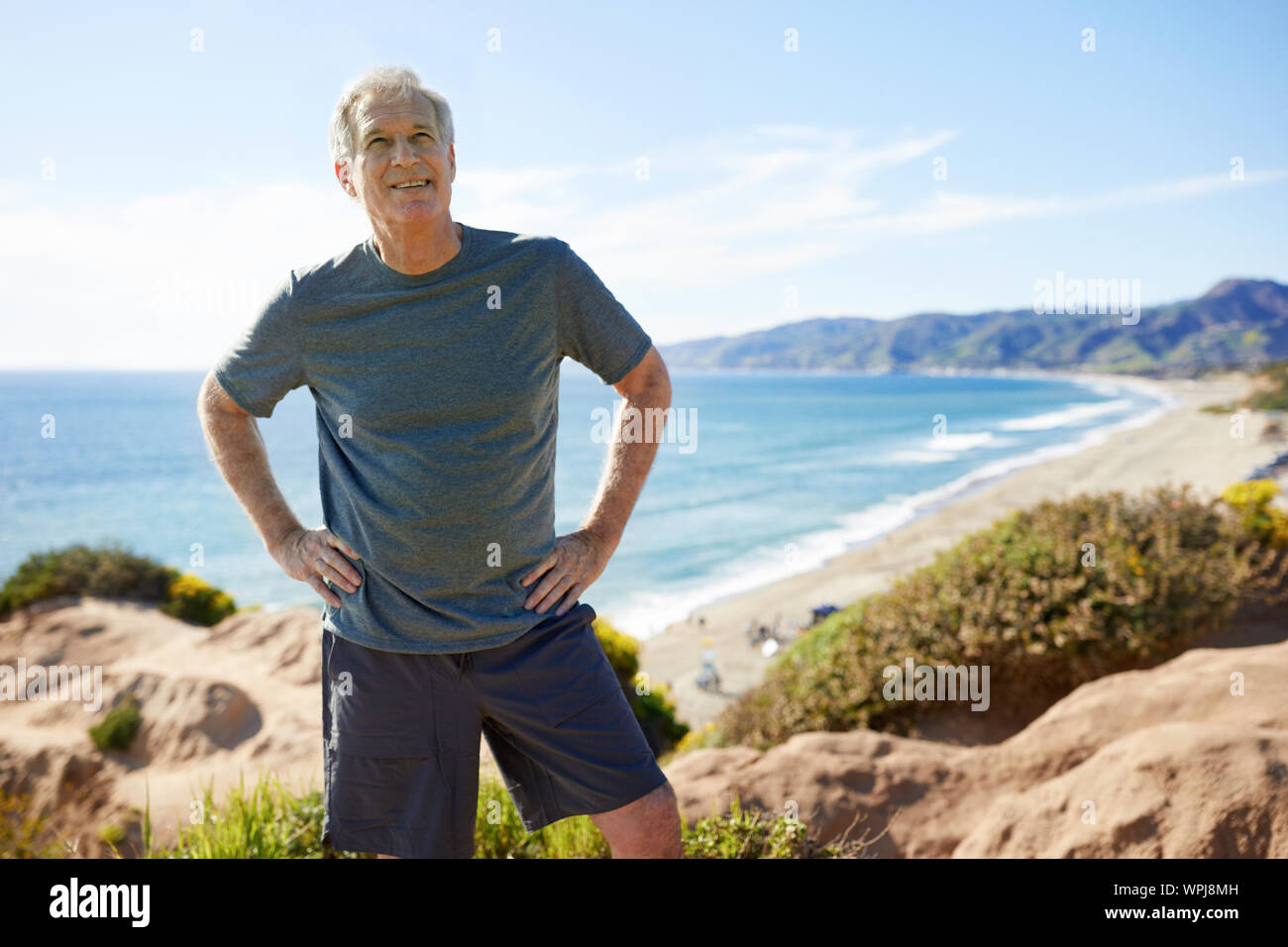 Lächelnd älterer Mann mit Händen auf den Hüften stehen auf Felsen am Strand gegen Sky Stockfoto