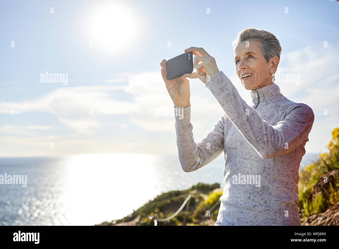 Lächelnd senior Frau Fotografieren durch Smart Phone, während gegen Himmel während der sonnigen Tag stehend Stockfoto