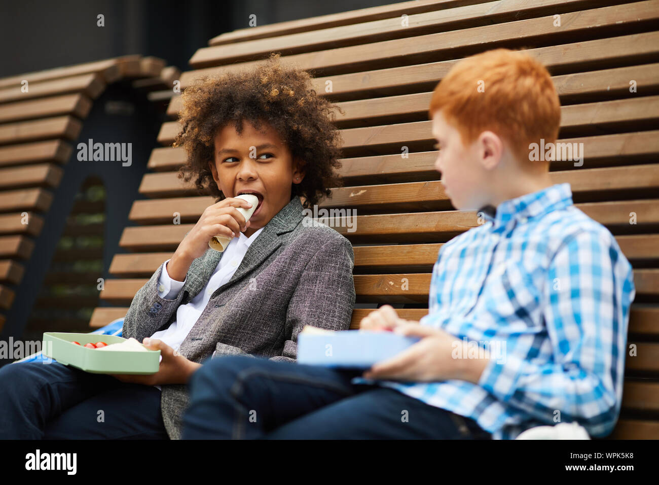 Afrikanische Schüler essen Sandwich zusammen mit seinem Freund, während Sie auf der Bank sitzen im Freien Stockfoto