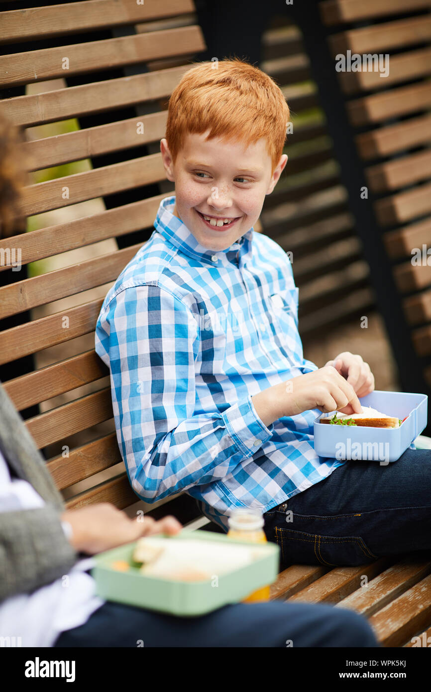 Rothaarige Junge in Freizeitkleidung auf der Bank sitzen Essen Sandwich und im Gespräch mit seinem Freund im Freien Stockfoto