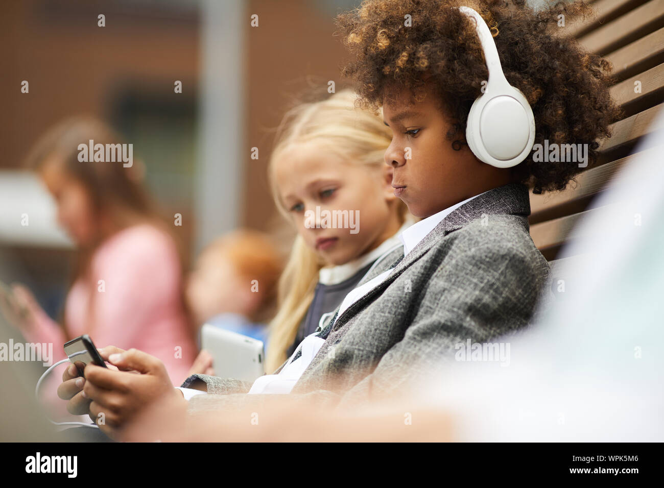 Afrikanischen Jungen mit dem lockigen Haar das Tragen von Kopfhörern und Spielen online Spiel auf sein Handy mit Mädchen sitzt in der Nähe von ihn auf die Bank im Freien Stockfoto