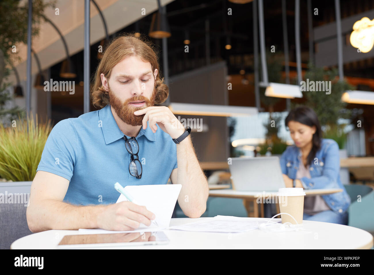 Ernsthafte bärtigen Geschäftsmann in Freizeitkleidung sitzen am Tisch und Schreiben seine Pläne für die Zukunft im Notepad mit Frau Arbeiten am Laptop Stockfoto