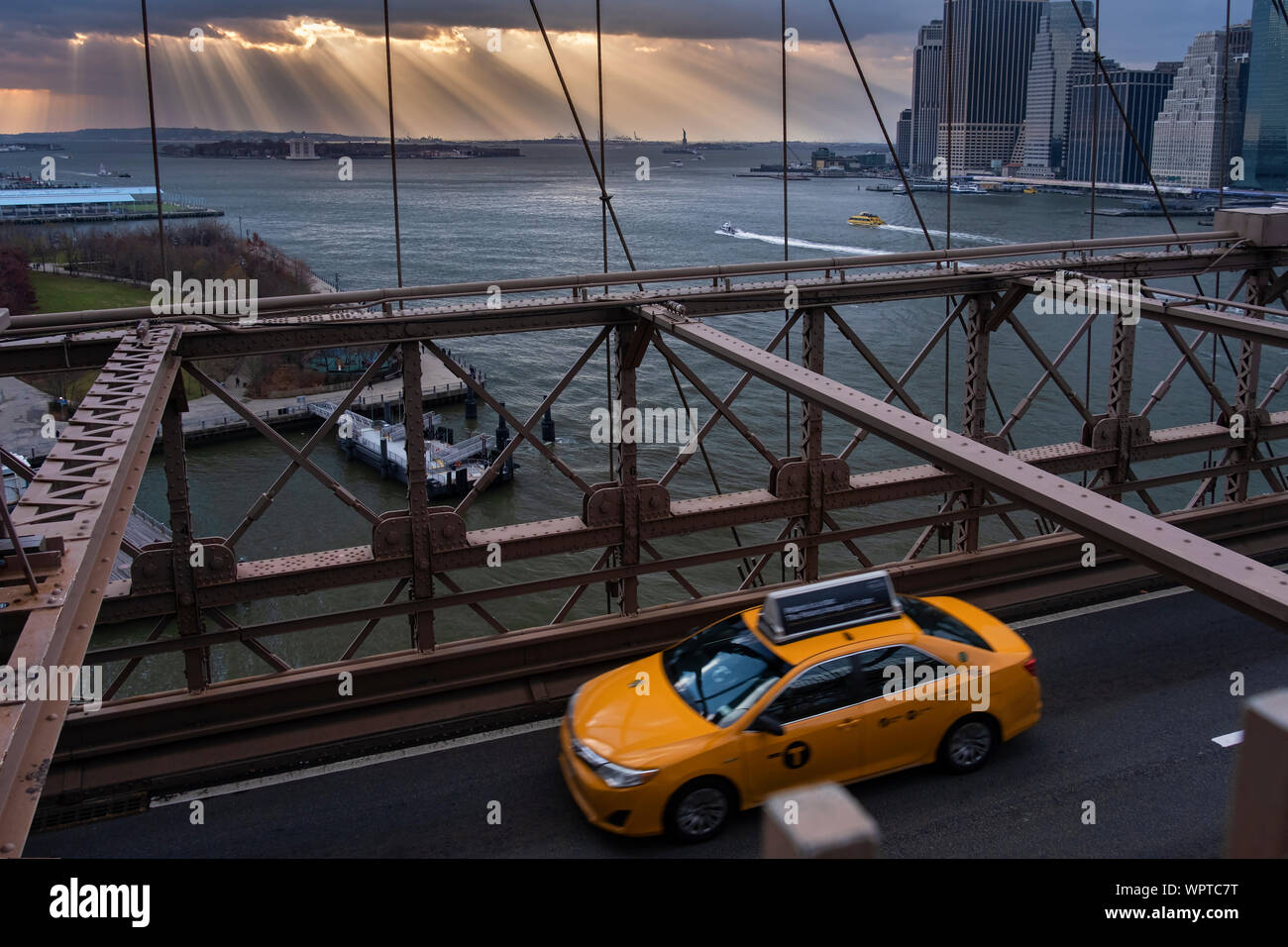 Yellow Cab überqueren Sie die Brooklyn Bridge mit Sonnenstrahlen über eine ferne Freiheitsstatue, New York, USA Stockfoto