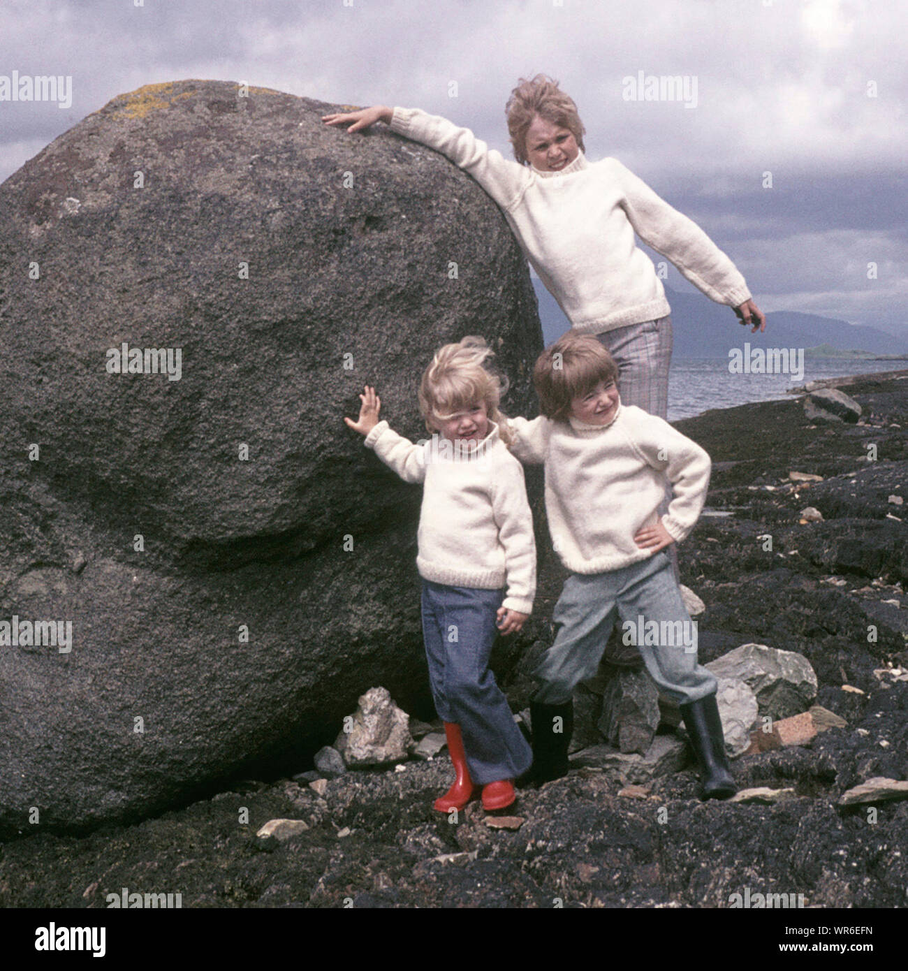Historisches Archivmodell 1970er veröffentlichte Familienurlaub in Schottland drei Kinder aus der gleichen Familie posieren in einem 70s passenden Aran-Modepullover und versuchten, den Felsblock neben dem kalten windigen loch zu schieben, wie wir in den schottischen Highlands in Großbritannien waren Stockfoto