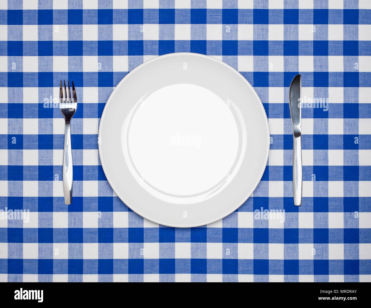 Ort, Einstellung, runde weiße Platte, Messer und Gabel, von oben auf blau kariertem Tischtuch Stockfoto