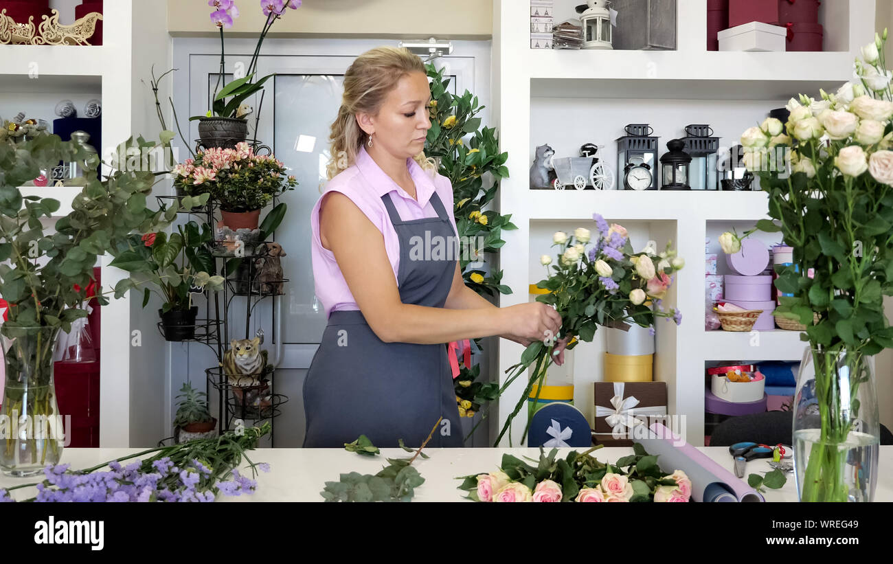Professionelle Floristen Frau schafft Schönen Blumenstrauß mit Rosen, Blätter und Gypsophila. Blonde Frau mittleren Alters in Uniform arbeitet im Blumengeschäft. Sie macht Haufen zarte Farben pink und gelb. Stockfoto