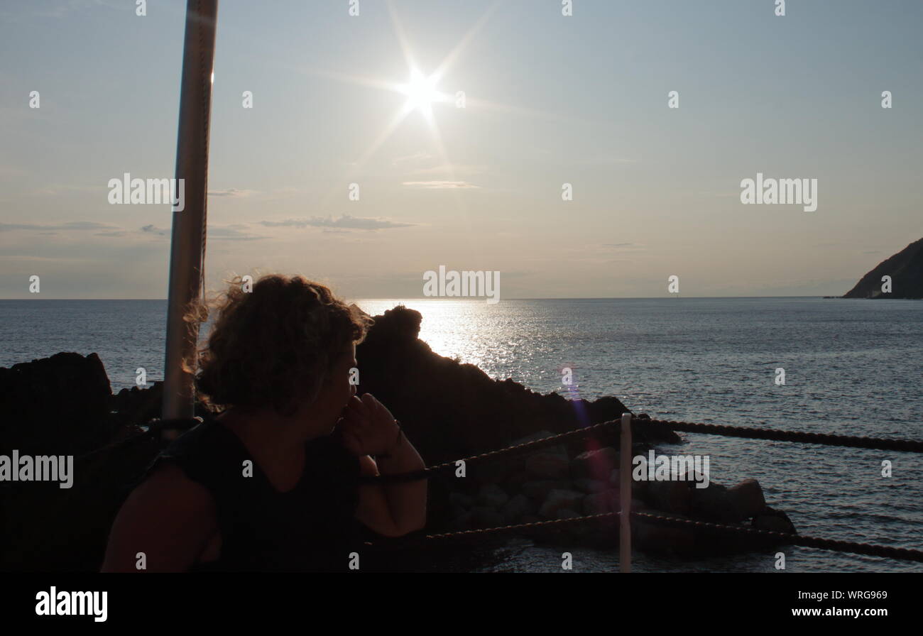 Eine caucasic blonde Frau mit einem Segelboot auf dem Horizont bei Sonnenuntergang am Strand bar Stockfoto