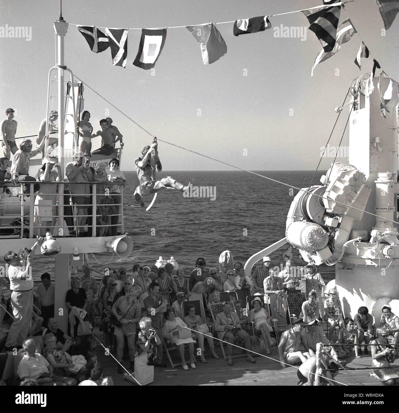 1960, historische, Spiel auf einem Kreuzfahrtschiff, Mitreisenden beobachten, wie einen männlichen Fahrgast ein Zip Line wird über das Deck des Schiffes auf dem Meer zu gehen. Stockfoto