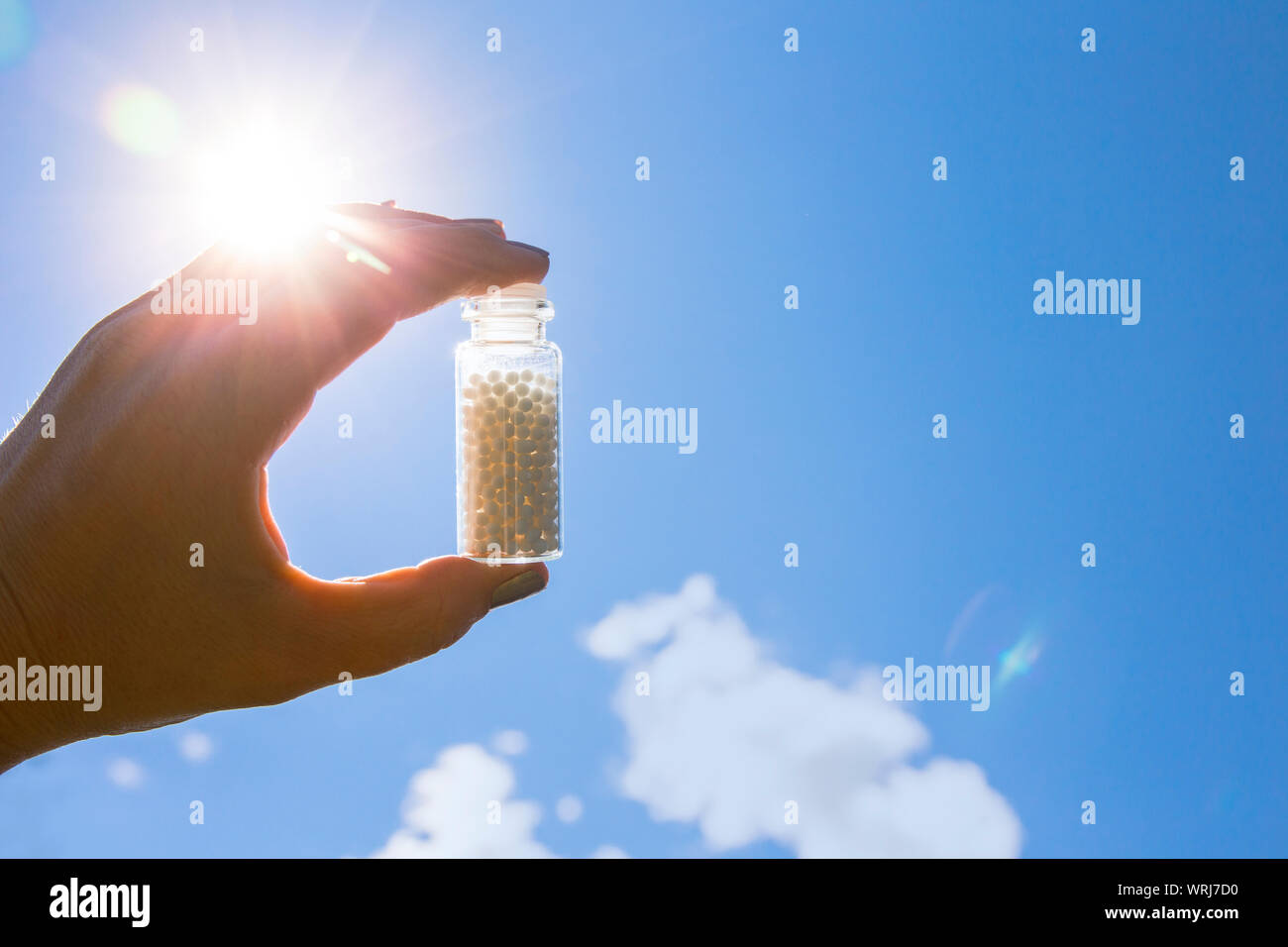 Selektiver Fokus auf Person hand Glas Glas voll mit kleinen weißen runden Homöopathie Pillen gegen den blauen Himmel Hintergrund. Stockfoto