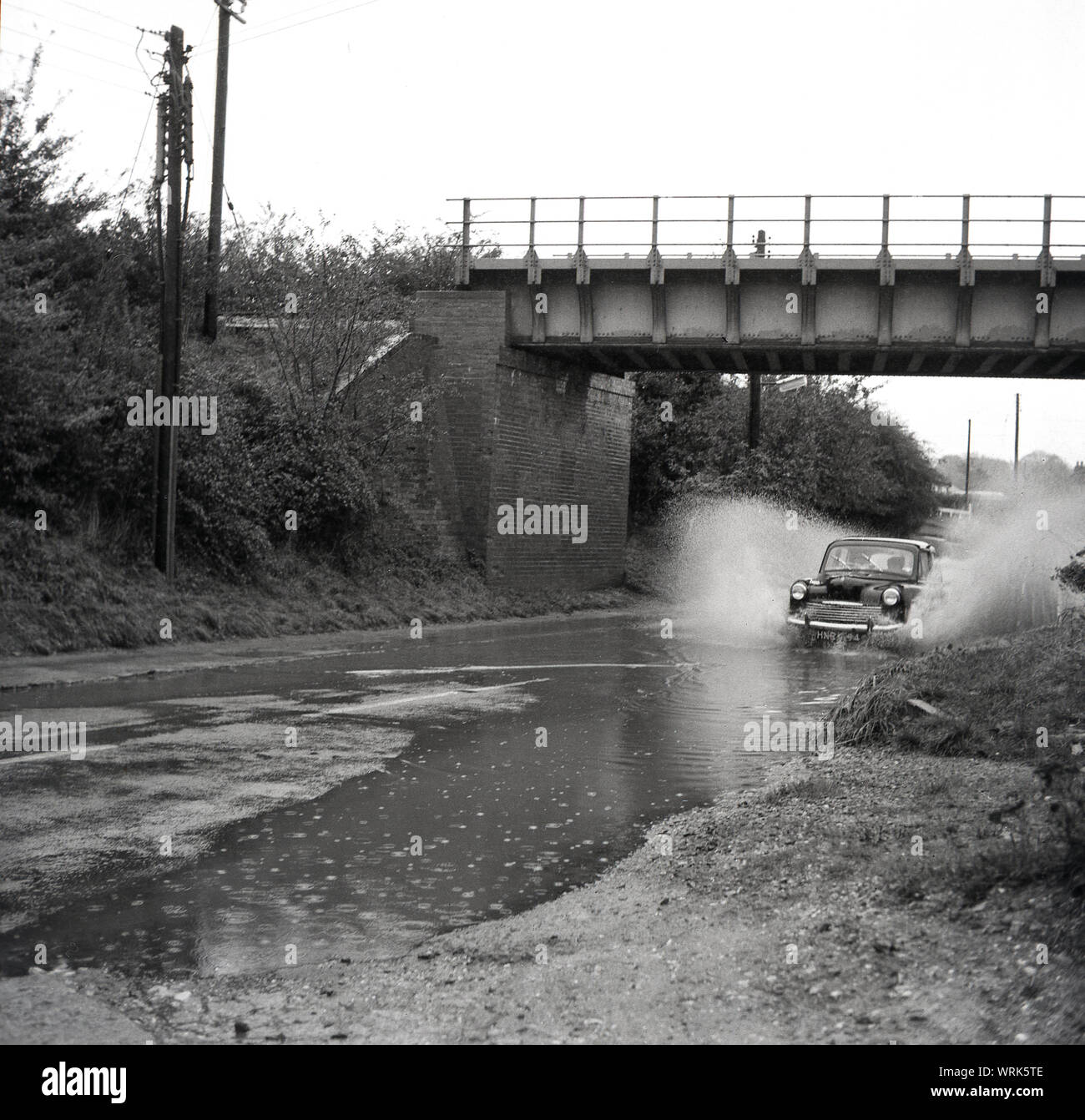 1967, historische, überflutete Straße, Auto der Ära unter einer Brücke durch einen großen Pool von Wasser auf einer Straße, wo die Kanalisation übergelaufen sind. Risborough, England, UK. Stockfoto