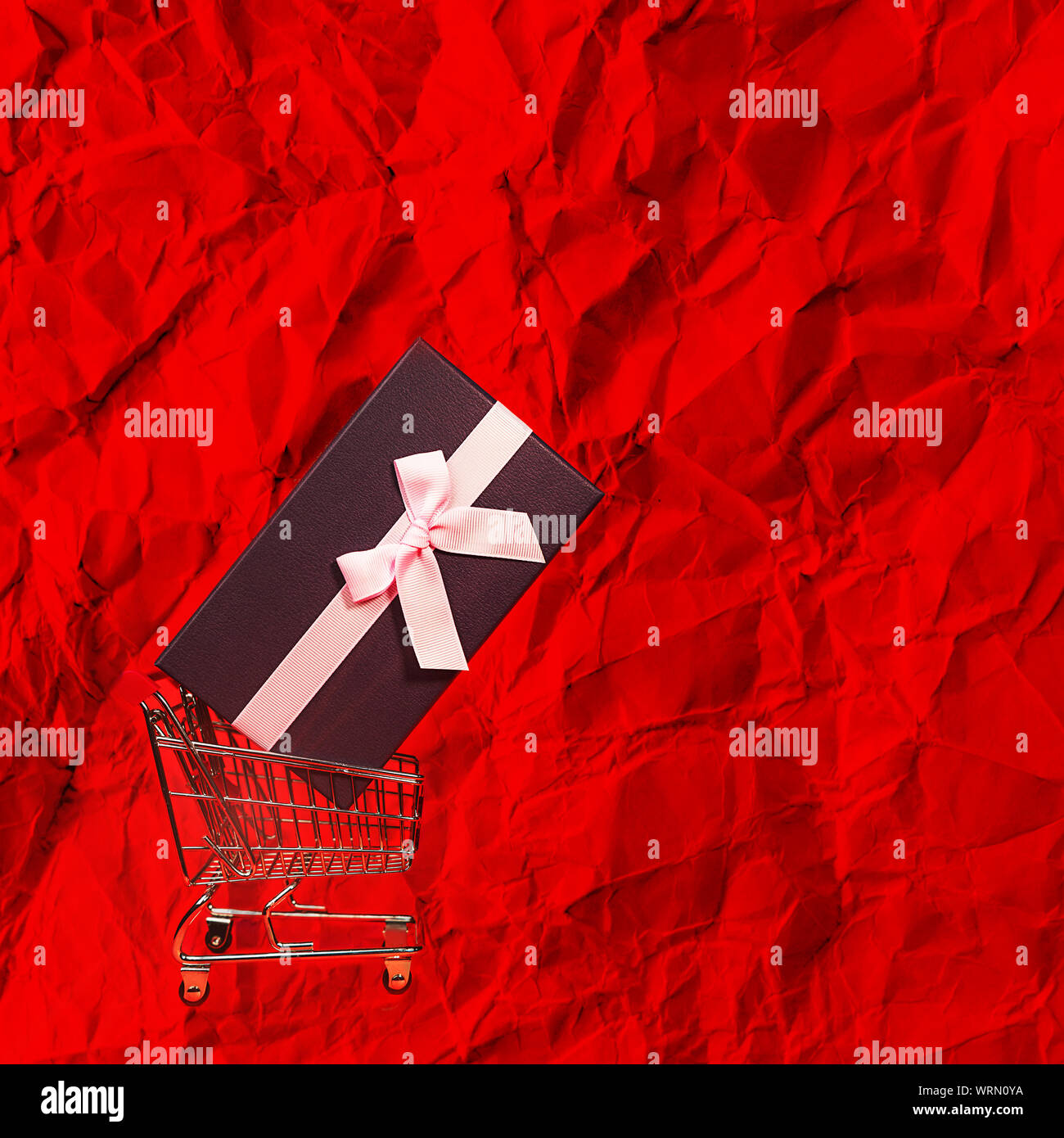 Urlaub Geschenk, Schwarzer Freitag Konzept mit Geschenkbox und rosa Schleife in einem Metall Einkaufswagen, Korb auf rote Farbe Geschenkpapier Hintergrund. Stockfoto