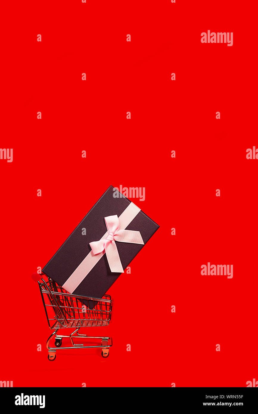 Urlaub Geschenk, Schwarzer Freitag Konzept mit Geschenkbox und rosa Schleife in einem Metall Einkaufswagen, Korb auf rote Farbe Hintergrund. Stockfoto