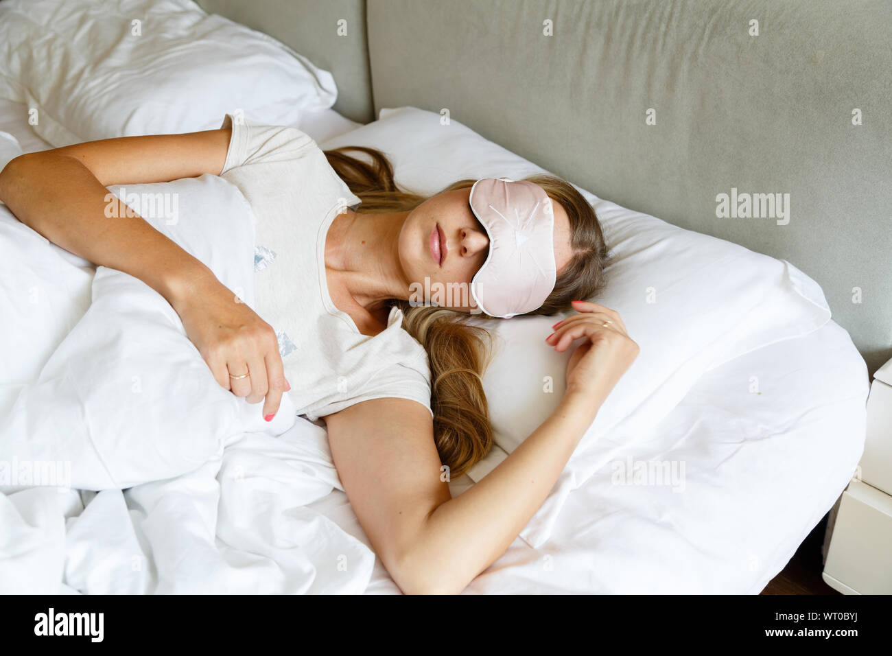 Junge Frau mit langen blonden Haaren schläft auf bequemes Bett in einer Maske zum Schlafen. Augenbinde auf Auge. Morgens zu Hause. Weiß Kissen und Decke Stockfoto