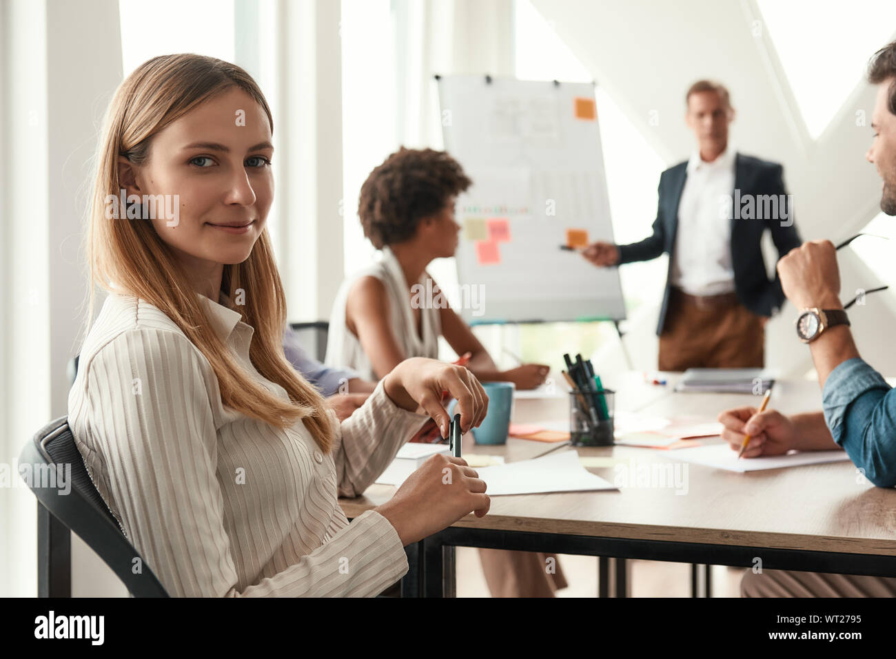 Junge Frau sucht zur Kamera und lächelnd, während Ihr Chef in der Nähe von Whiteboard und etwas diskutieren mit Team. Teamarbeit. Konferenz Stockfoto