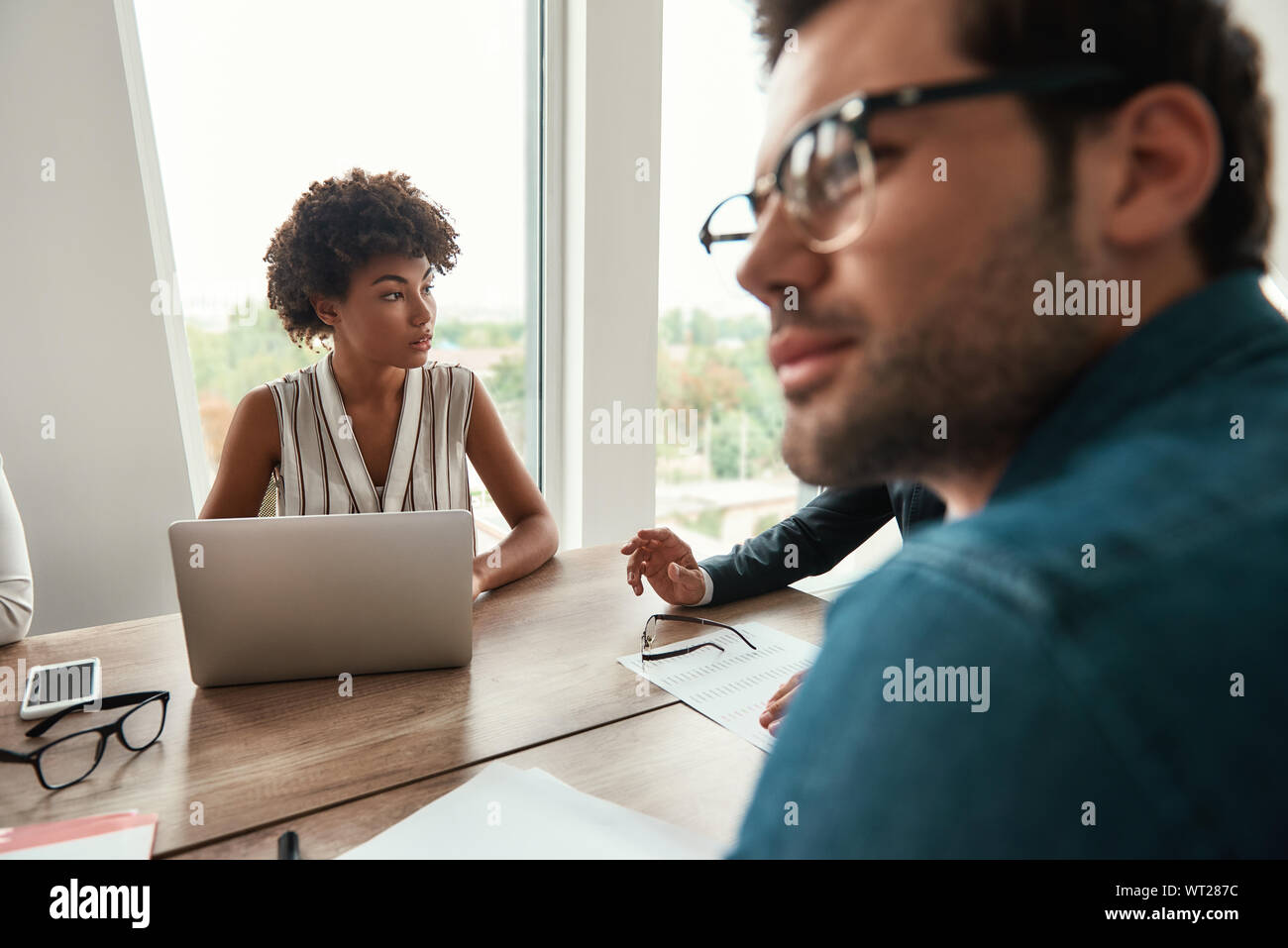 Zusammen zu arbeiten. Junge und schöne afro-amerikanische Frau, die Arbeiten am Laptop und etwas diskutieren mit Kollegen, während im modernen Büro zu sitzen. Brainstorming. Business Meeting Stockfoto