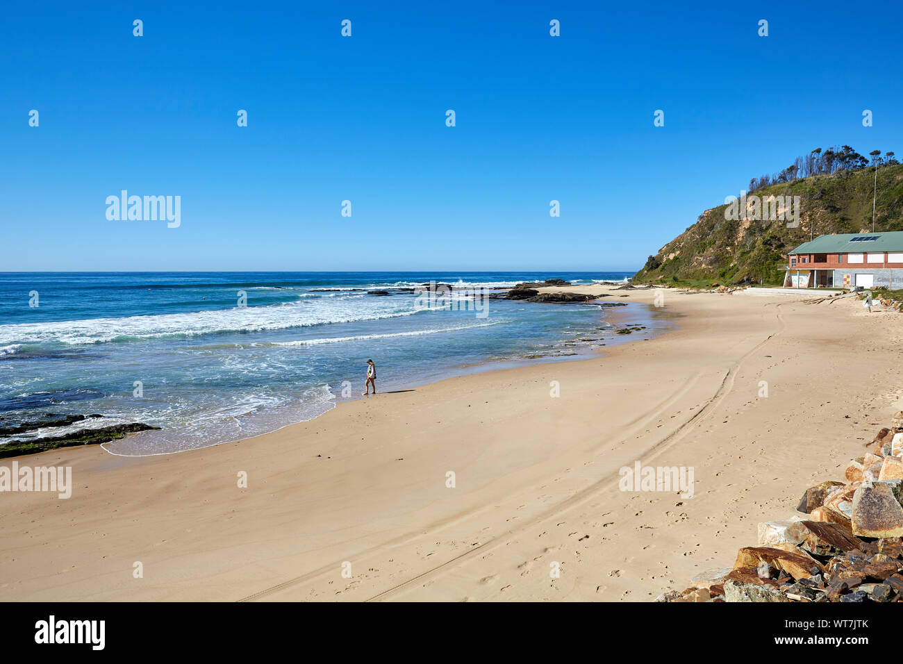 Eine Person, die in der Ferne allein zu Fuß entlang der Ufer von einem Strand an einem sonnigen, wolkenlosen Herbsttag auf der Küste von Australien Stockfoto