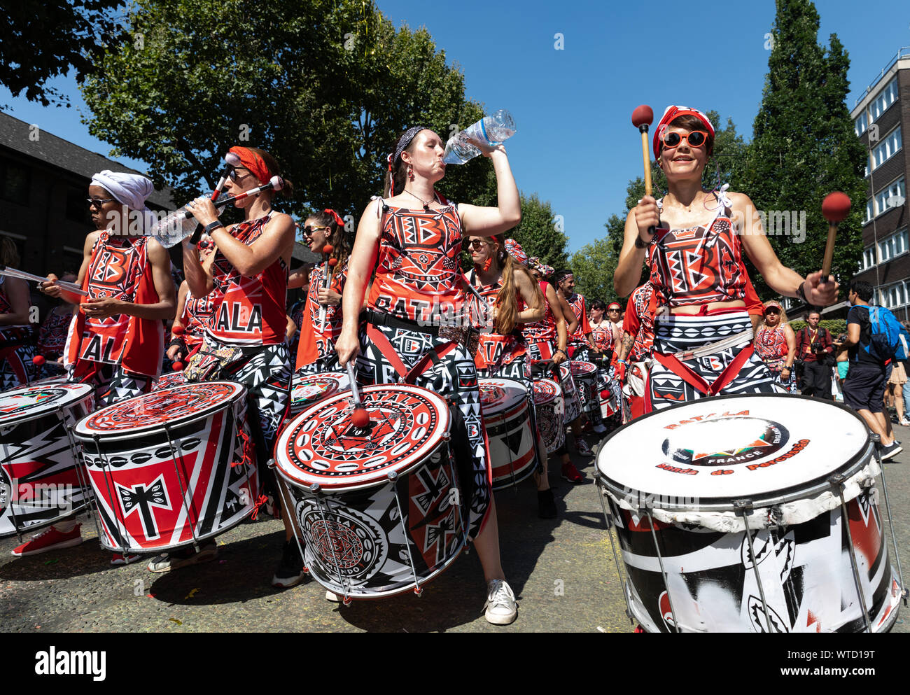 Ein drumming Band in Rot, Weiß und Schwarz gedruckt Kostümen trinken Flaschen Wasser, wie Sie an Notting Hill Carnival am 26. August 2019 durchführen. Stockfoto