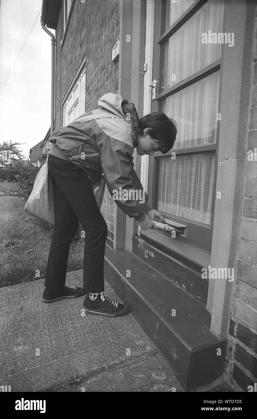 1980 s, historischen, ein paperboy auf die Bereitstellung einer Abendzeitung durch einen Briefkastenschlitz biegen. Nach der Schule, ein paperboy mit Ranzen, seine Nachmittag oder frühen Abend Papier runde Tun auf einer Wohnsiedlung wurde ein alltäglicher Anblick in England, Großbritannien. Oft erster Job eines jungen Menschen, es gab Ihnen Ihre erste Erfahrung mit der Arbeit und ein kleines Einkommen, aber die verlieren viele Abend gedruckte Zeitungen hat ihre Zahl rapide abnehmen aus früheren Jahrzehnten gesehen. Stockfoto