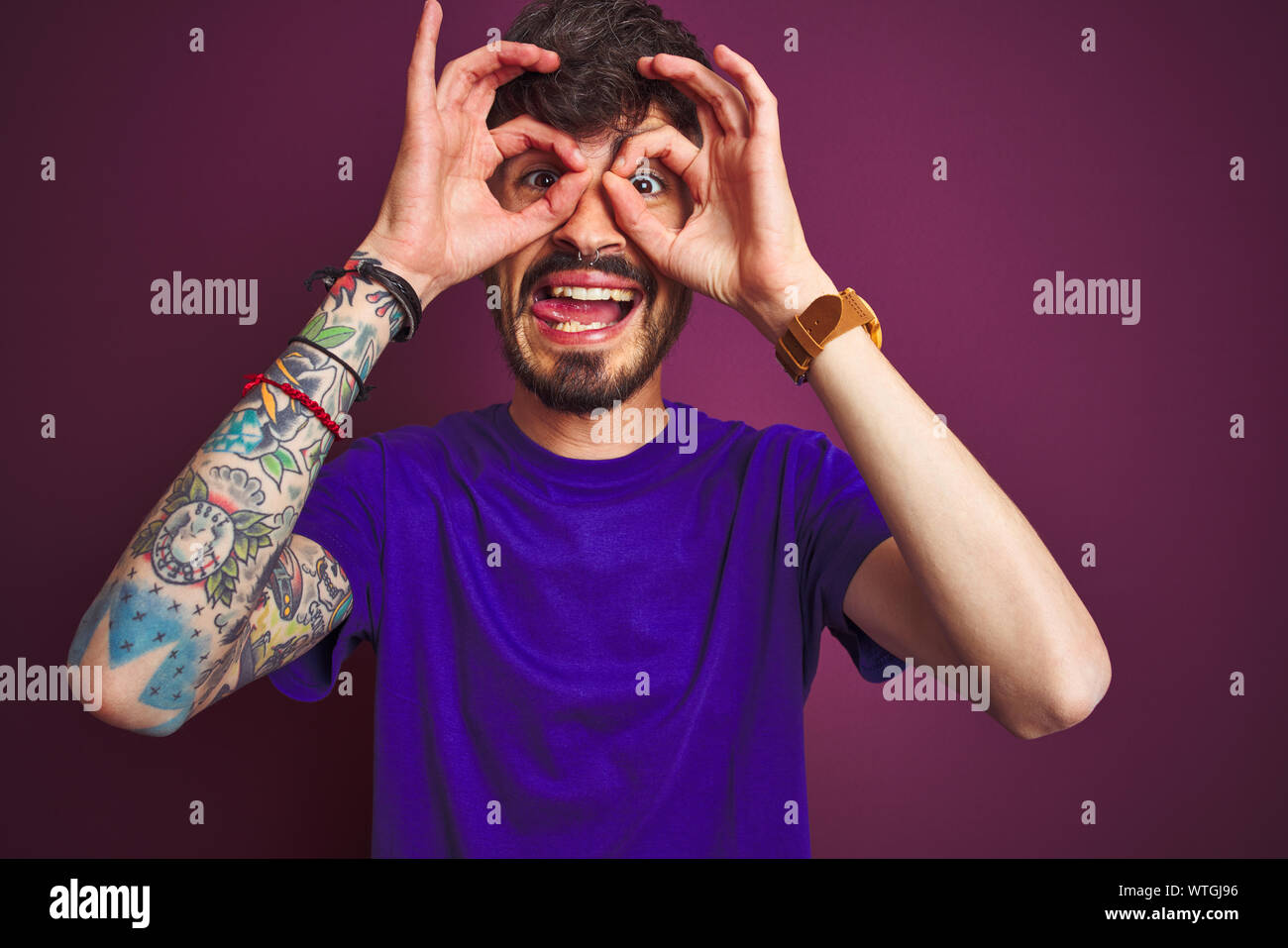 Junger Mann mit Tattoo tragende t-shirt stehend über isolierte lila  Hintergrund tun ok Geste wie Fernglas Zunge heraus, Augen, thro  Stockfotografie - Alamy