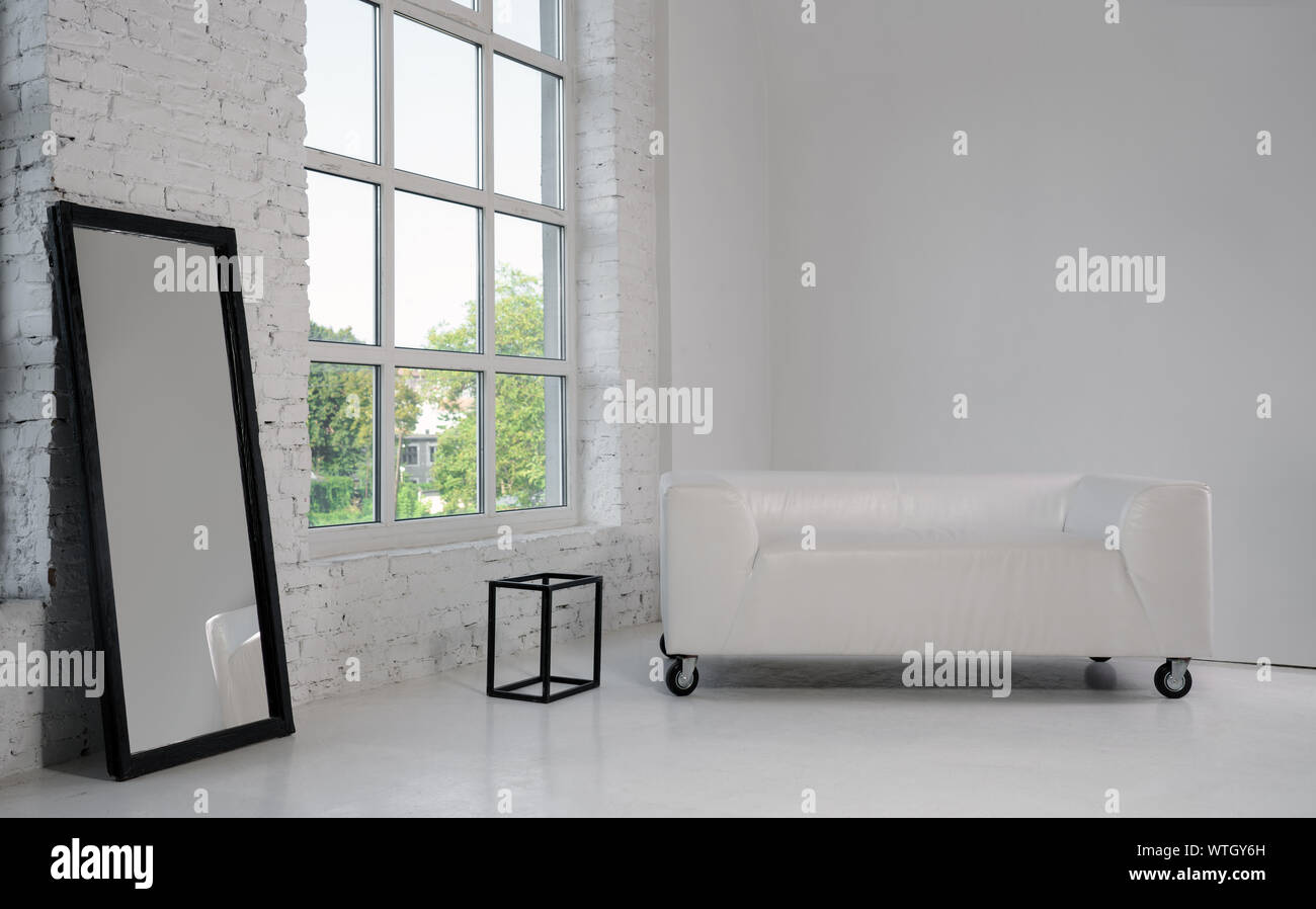 Modernes weißes Sofa und große schwarz gerahmten Spiegel in weißen minimalistischen Zimmer mit grossem Fenster Stockfoto