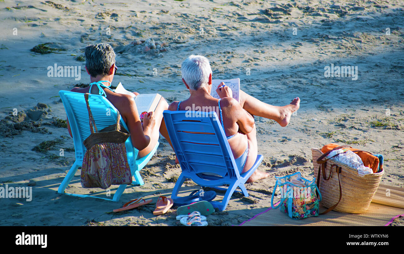 Alicante, Spanien, 25. August 2019: Zwei ältere Frauen am Strand Kreuzworträtsel lösen und lesen Sie beim Bräunen sitzt. Sommer für Rentner oder Pensionäre. Stockfoto