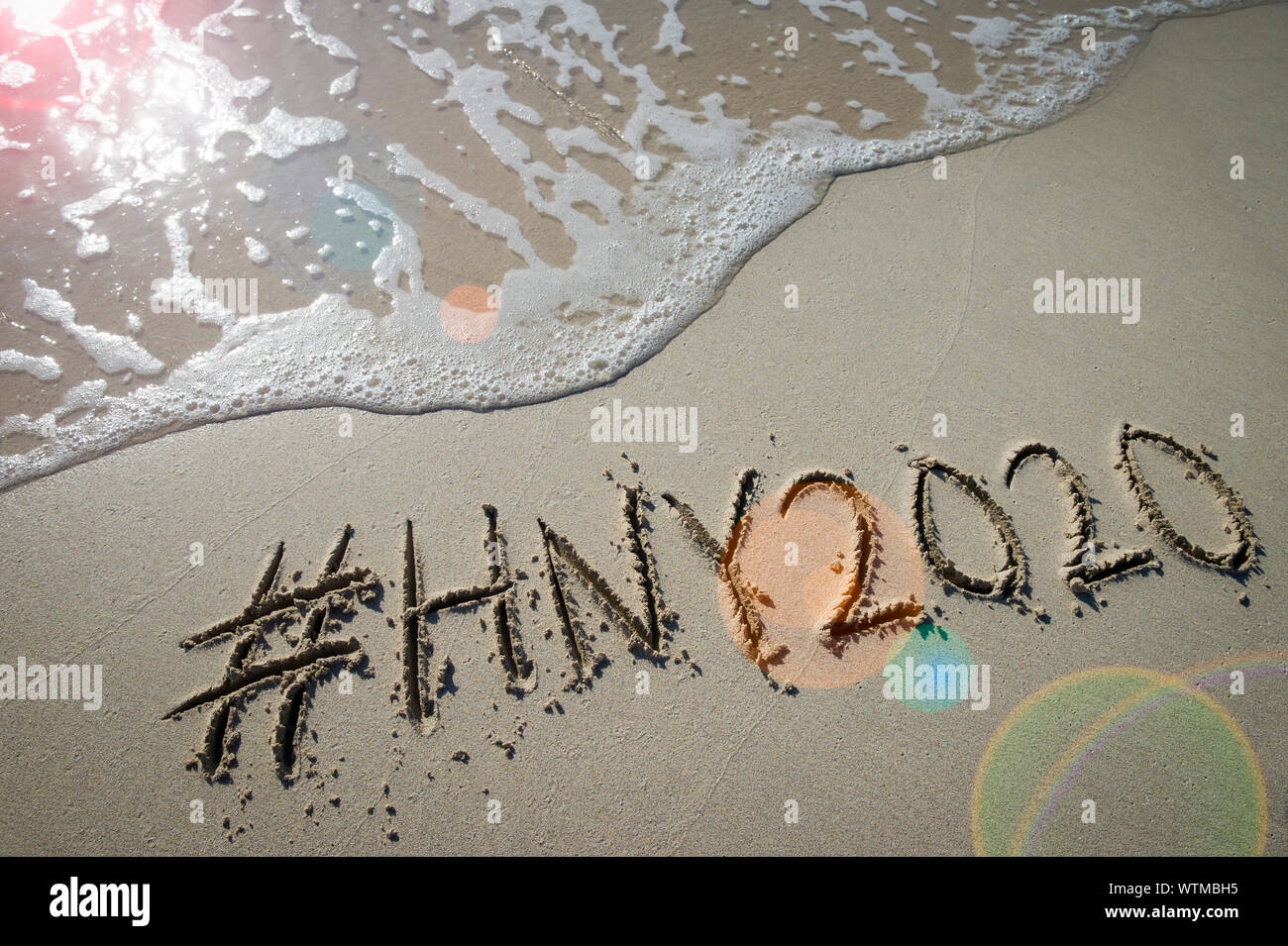 Modernes neues Jahr Nachricht mit social media Hashtag für 2020 handschriftlich in sanften Sand mit Sunrise lens flare über entgegenkommende Welle auf den Strand Stockfoto