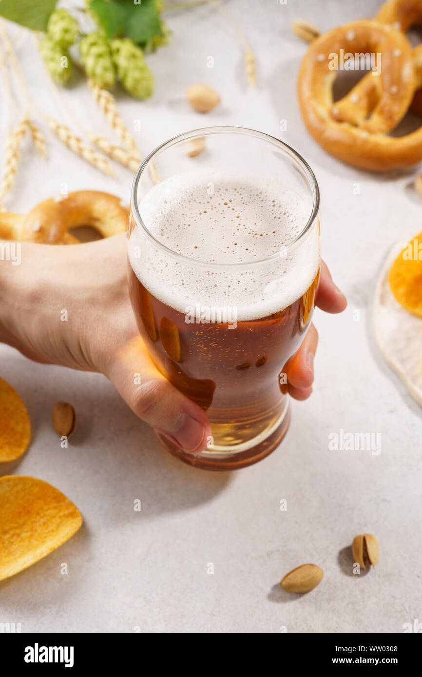 Mann hand mit einem Glas helles Bier. Verschiedene Snacks im Hintergrund - Brezeln, Chips und Nüsse. Stockfoto