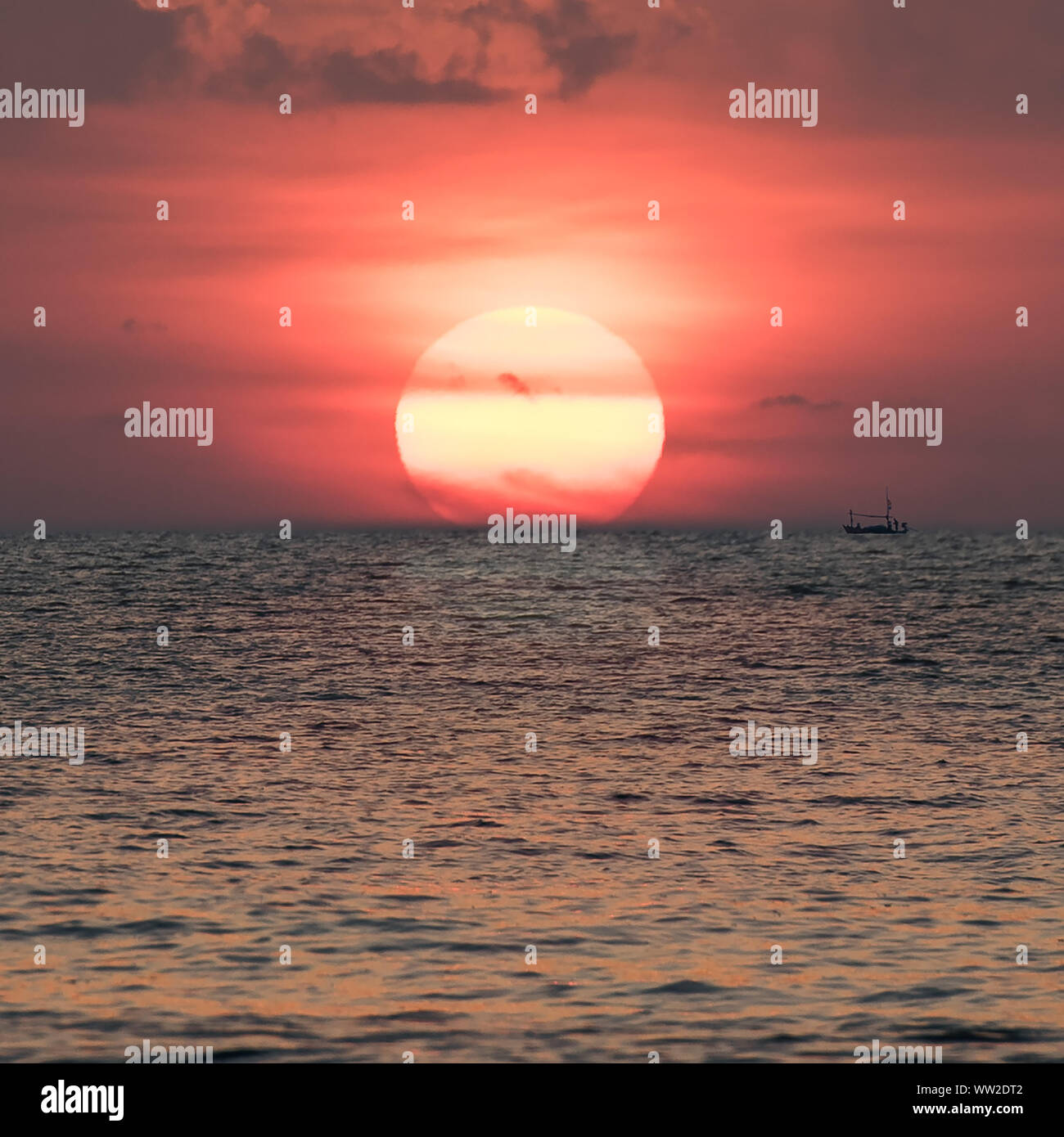 Ein kleines Fischerboot mit einer Spiegelung der Sonne mitten im Meer bei Sonnenuntergang. Stockfoto