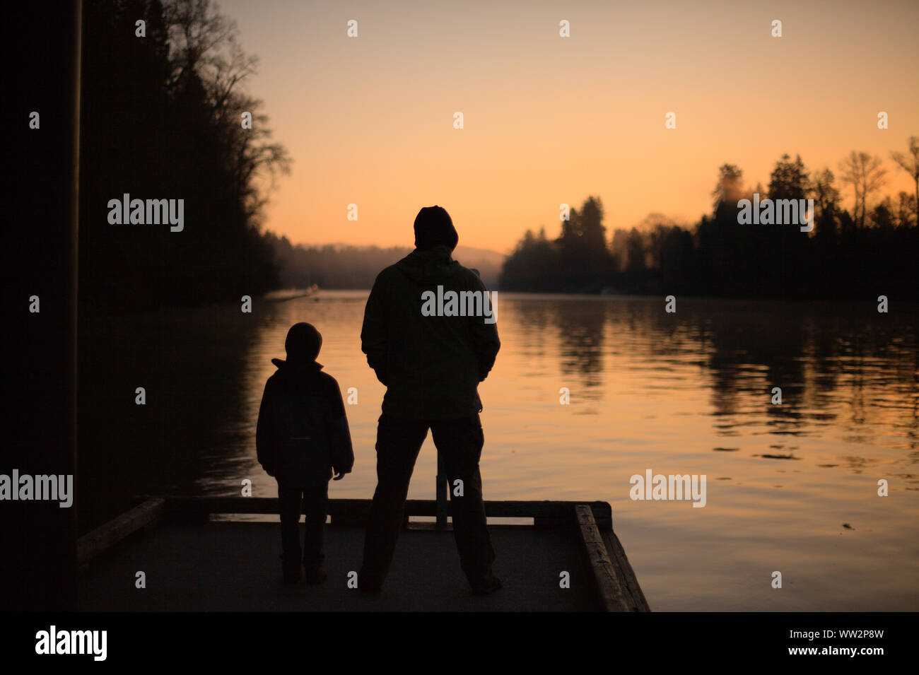 Ein Mann und Kind dunkle Silhouette auf einem Dock beobachten die Orange Sonnenaufgang auf Willammette River, in den kalten orange Frühling Morgen Licht der Wilsonville, ODER Stockfoto