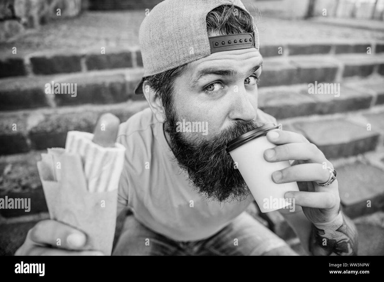 Trinken Sie etwas anders. Kaukasische hipster Trinken Essen trinken mit Hot Dog. Man nimmt einen Schluck frischen Kaffee trinken im verfügbaren cup Bärtigen. Ernährung Essen und Trinken. Stockfoto