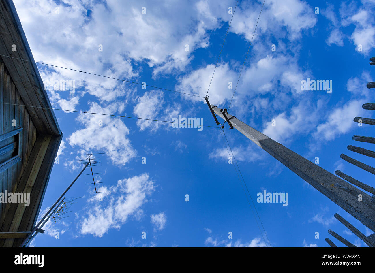Konkrete Pol mit elektrischen Leitungen neben einem hölzernen Zaun und ein Teil des Daches von einem Haus im Dorf im Hintergrund ein blauer Himmel mit Wolken Stockfoto
