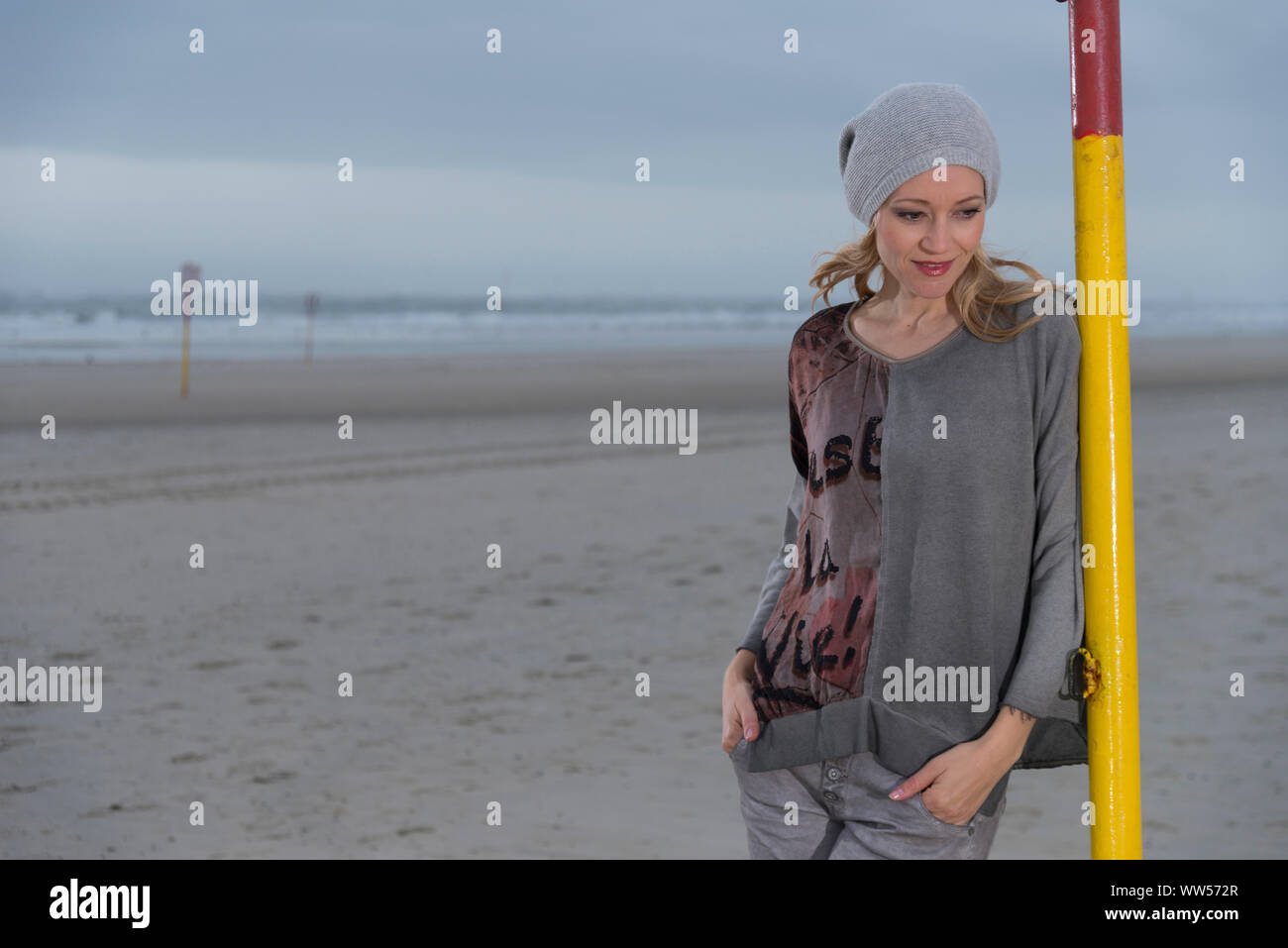 Blonde Frau mit grauer Kappe schiefen auf gelbem Schild Säule am Strand Stockfoto