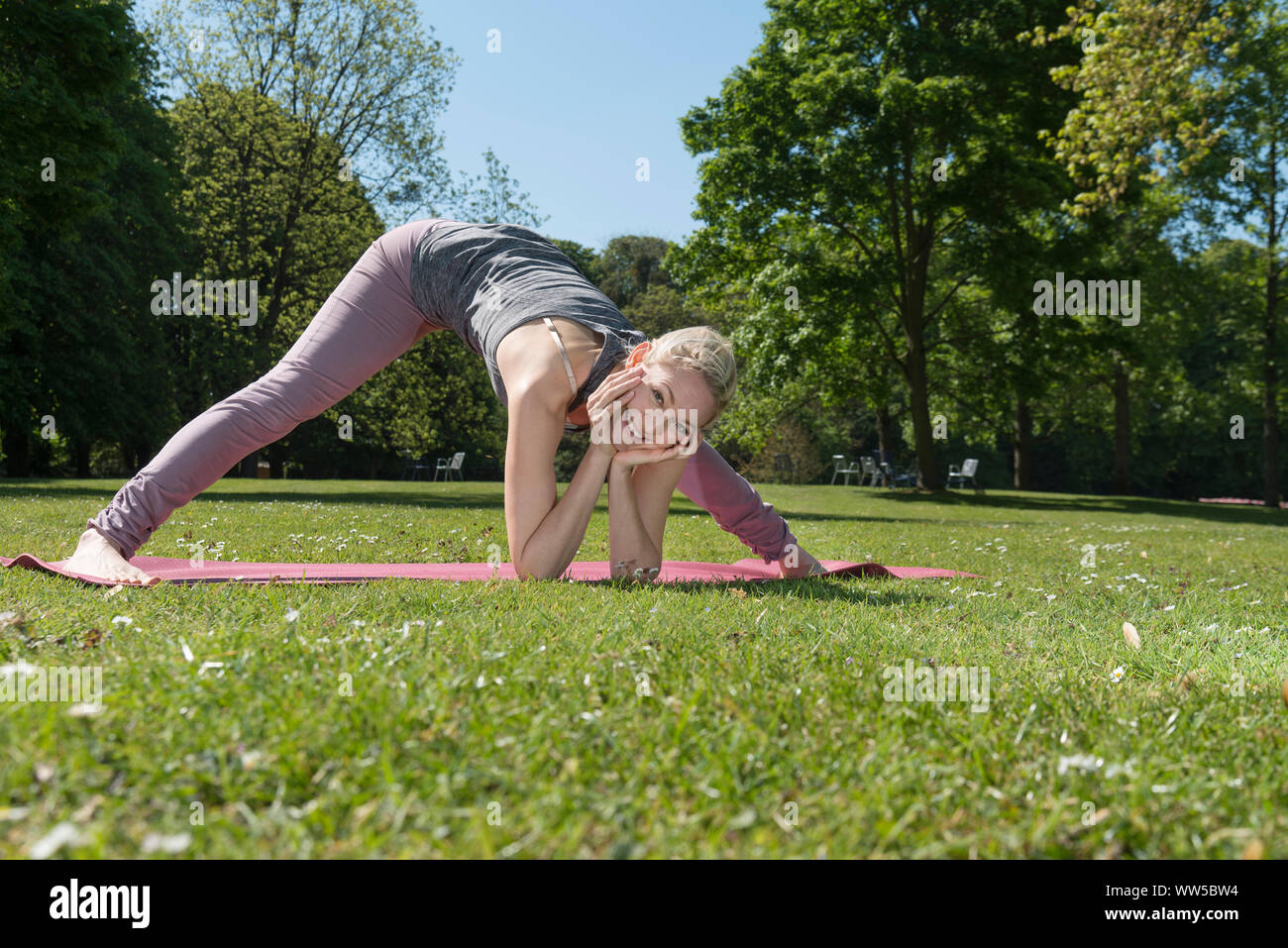 Frau in Trainingskleidung auf rosa matt im Park, Yoga, Gymnastik, Bücken, Kopf in den Händen, lächelnd, bei Camera suchen Stockfoto